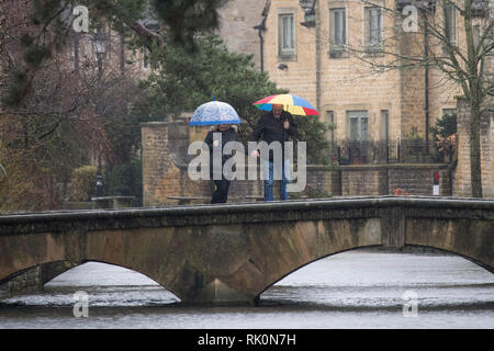 Tourists with their umbrellas as they cross a bridge during a rain shower on a wet and windy day in Bourton-on-the-Water in Gloucestershire. 08.02.19. Stock Photo