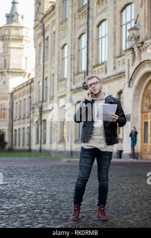 Guy in casual clothes holding papers and talking on the phone, standing next to the university Stock Photo