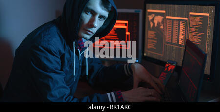 Male in the hood looking in the camera. Hacker attacks and online fraud in the dark on the screen background in neon light. The concept of cyber secur Stock Photo
