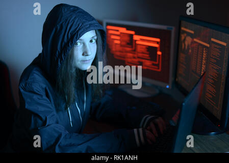 Girl in the hood looking in the camera. Hacker attacks and online fraud on the screen background in neon light. The concept of cyber security Stock Photo