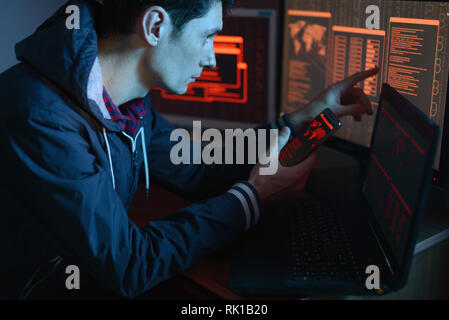 The male hacker in the hood is pointing his finger at the display indicating the location of the cyberattack and hacking data on the screen background Stock Photo