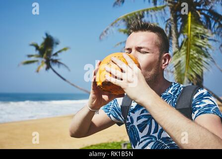 Thirsty young man drinking coconut water. Relaxation on the sand beach. Sri Lanka Stock Photo