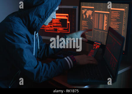 The male hacker in the hood is pointing his finger at the display indicating the location of the cyberattack and hacking data on the screen background Stock Photo