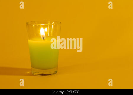 Small glass container containing a candle of colored color on a yellow background. Candle lit. Stock Photo