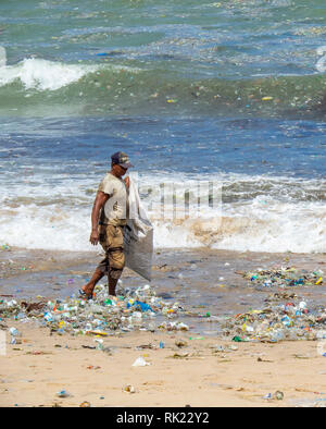 Pollution, lone man picking up plastic bottles, cups, straws and other litter washed up on the beach at Jimbaran Bay, Bali Indonesia.. Stock Photo