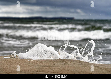 A small wave breaks on the Baltic Sea beach in front of a contrasting sky, the spray and single drops of water stand out against the background - Loca Stock Photo
