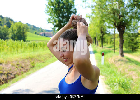 Woman tying hair in ponytail getting ready for exercising. Beautiful young sporty woman attaching her long hair in park. Smiling woman tightening hair Stock Photo