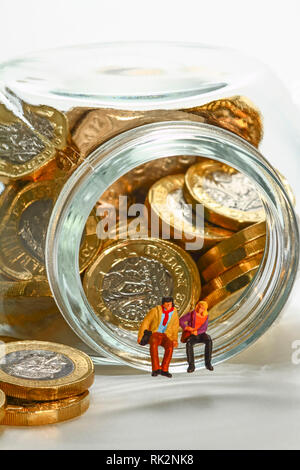 Conceptual diorama image of miniature figures sat on a glass jar filled with pound coins Stock Photo