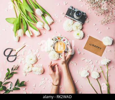 Flat-lay of female hands holding cup of espresso coffee, scissors, film camera, white tulips and buttercup flowers and sign happy over pink background Stock Photo