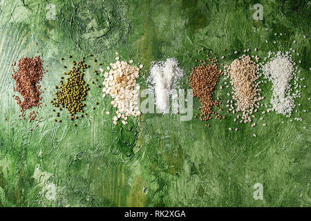 Variety of raw uncooked grains superfood cereal linen seeds, sesame, mung bean, wheat, buckwheat, oatmeal, coconut, rice over green texture background Stock Photo