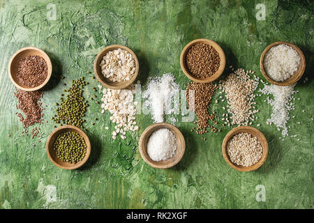 Variety of raw uncooked grains superfood cereal linen seeds, sesame, mung bean, wheat, buckwheat, oatmeal, coconut, rice in wooden bowls over green te Stock Photo