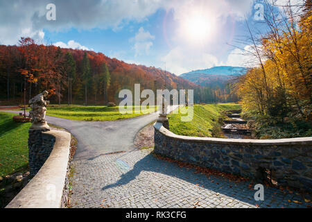 park among mountain in autumn. trees in colorful foliage, vivid grassy green lawns. walking path to the bridge through creek. wonderful sunny weather Stock Photo