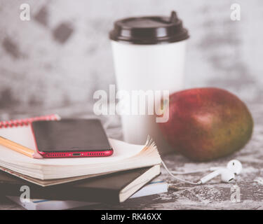 Working or education on the go. Coffee cup, mango fruit, red smartphone and stack of notepads on grey stone table. Copy space. Stock Photo