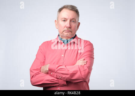 Serious senior man with folded arms and a deadpan expression posing on grey wall Stock Photo