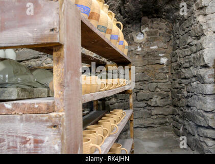 handcrafted clay pots and cups sitting on display on wood shelves in an underground room with old brick walls. Stock Photo