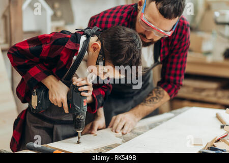 Handsome father and his little pre-teen son dressed in checkered shirts and wearing protective glasses working with drill perforating wood plank at wo Stock Photo