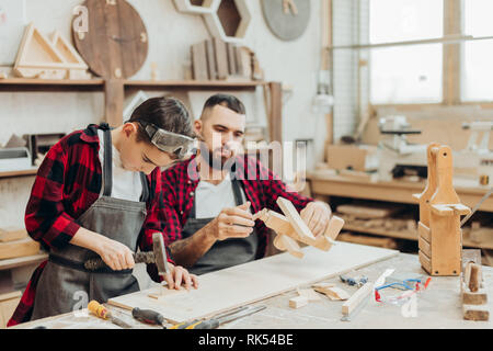 Carpenter building a wooden airplan together with his kid. A little son is participating actively in hand made process. Happy fatherhood and DIY conce Stock Photo