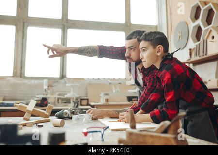 family, carpentry, woodwork and people concept - Carpenter pointing at something and little son looks at it at workshop Stock Photo