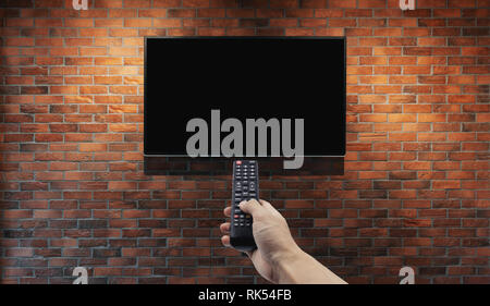 Television on brick wall with hand using remote control Stock Photo