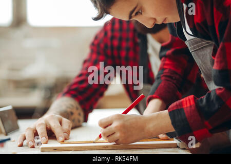 Young father is keen on making DIY wooden toy together with his son on carpentary masterclass in woodworking studio. Dad is helping his son to mark up Stock Photo