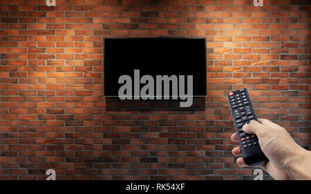 Television on brick wall with hand using remote control Stock Photo