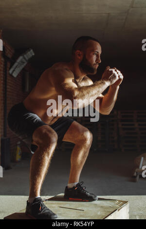 Determined young man jumping onto a box as part of exercise routine. Man doing box jump in the gym. Athlete is performing box jumps Stock Photo