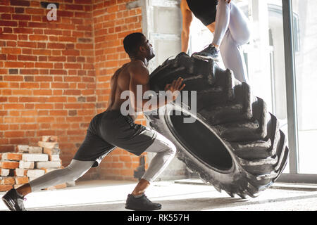Young muscular african man flipping a tire with a woman on it, working out with weighting Stock Photo