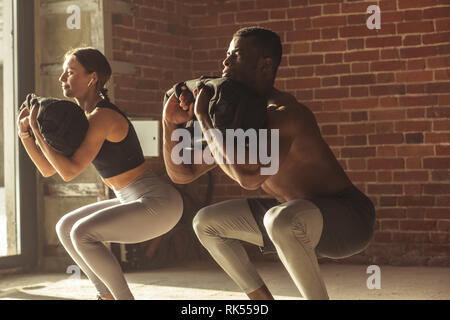 Young mixed-race fit couple doing squats exercise, with heavy sandbag in gym. A crossfit sand bag is advanced level for athletes of various sports, su Stock Photo