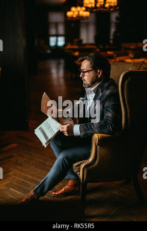 Adult man in his fifties, successful entrepreneur in spectacles and suit reading newspaper while sitting in luxury genuine leather arm-chair in dark r Stock Photo