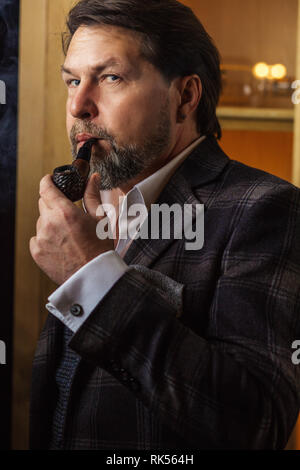 Experienced professional Chief executive, dressed in tailored suit, is puffing pipe, pondering over development strategy of company, making serious bu Stock Photo