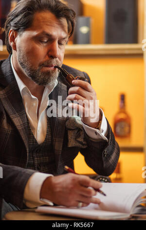 Talanted bearded writer in fashionable clothing is making notes in diary or notebook, sitting at restaurant and enjoying tobacco pipe Stock Photo