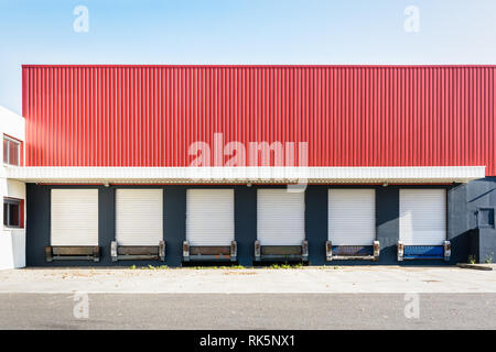 Front view of six truck loading docks at a warehouse with white roller shutter doors closed under a red corrugated iron siding. Stock Photo
