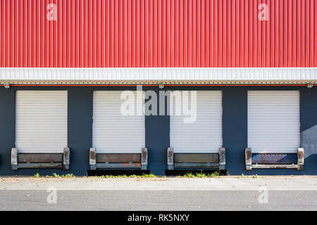 Front view of four truck loading docks at a warehouse with white roller shutter doors closed under a red corrugated iron siding. Stock Photo