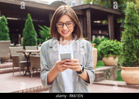 Business casual. Young lady in eyeglasses standing on the city street using app on smartphone happy Stock Photo