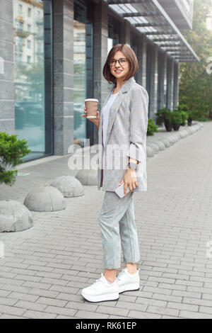 Business casual. Young lady in eyeglasses standing on the city street drinking coffee looking camera joyful full body shot Stock Photo