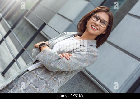 Business casual. Young lady in eyeglasses standing on the city street crossed arms looking aside happy Stock Photo