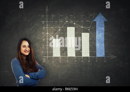 Pretty woman analyst holding arms crossed in front of a huge blackboard with drawn a increasing graph with arrows going up. Business industries profit Stock Photo