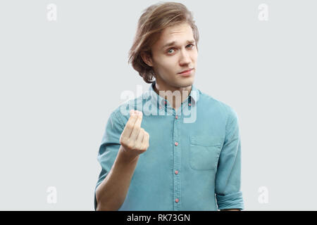 Portrait of serious handsome long haired blonde young man in blue casual shirt standing with money hand gesture and asking for more. indoor studio sho Stock Photo