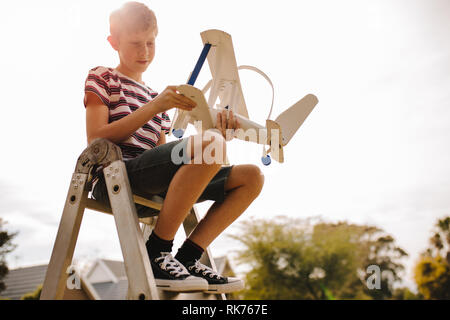 Boy sitting on top of a ladder and adjusting the toy airplane wing. Boy with toy aeroplane outdoors. Stock Photo