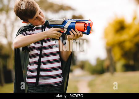 Young boys wearing a cape playing with a toy gun outdoors. Boy with cape pretending to a superhero. Stock Photo