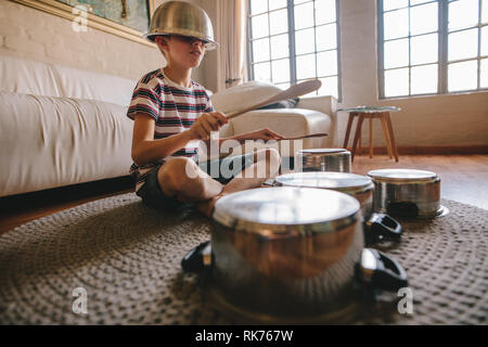 Boy playing drums on kitchenware at home. Boy pretending to be a drummer sitting on the living room floor and playing on utensils while wearing a bowl Stock Photo