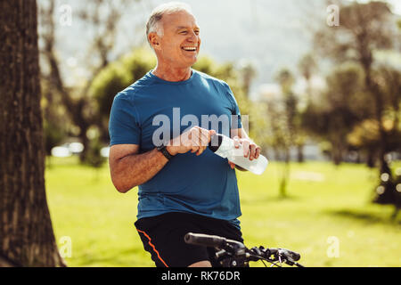 Senior man in fitness wear drinking water sitting on his bicycle. Cheerful senior fitness person taking a break during cycling in a park. Stock Photo