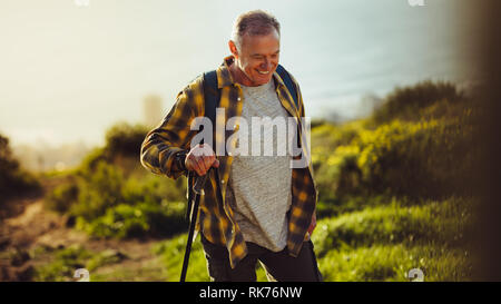 Cheerful man walking up a hill with the help of trekking pole. Man enjoying trekking up the hill on a sunny day. Stock Photo