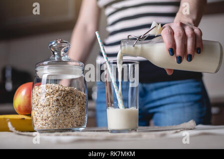 A woman pours organic oat milk from a bottle into a glass on a table in the kitchen. Diet healthy vegetarian product Stock Photo