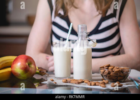 Woman drinking organic almond milk holding a glass in her hand in the kitchen. Bottle and fruit on the table. Healthy vegetarian product Stock Photo