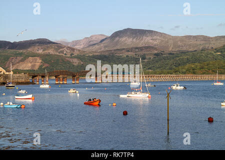 Boats moored on the Mawddach Estuary near Barmouth, Wales, with a passenger train crossing Barmouth Bridge and mountains in the background. Stock Photo