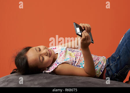 Kid smiling on a beanbag holding phone. Asian girl holding cellphone and being happy. Stock Photo