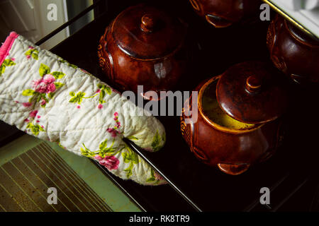 Woman taking out of oven stewed or baked potatoes in brown ceramic pots. Stock Photo