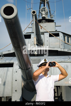 Captain of warship standing beside cannons and watching the surroundings. A navy officer in white uniform looking with binocular aboard a battleship. Stock Photo