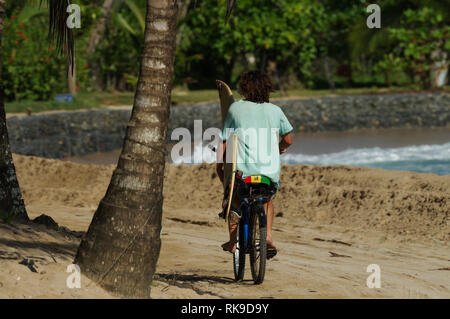 Young man on riding a bike along the beach bike carrying a surf board - Bocas Del Toro, Panama Stock Photo